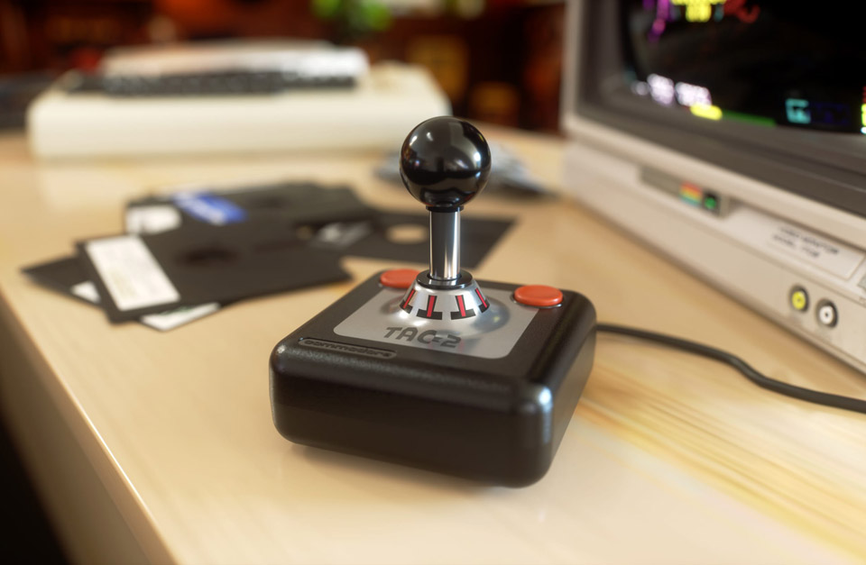 Le joystick TAC-2 dans son coloris noir "Knight Black" classique, avec ses boutons rouges, posé sur un bureau devant un moniteur et des disquettes souples