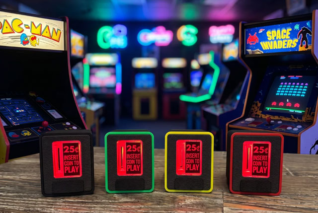 Quatre monnayeurs aux contours noirs, verts, jaunes et rouges, avec écrit en rouge "25 cents - Insert Coin to Play" près de la fente, le tout sur une table devant une salle d'arcade